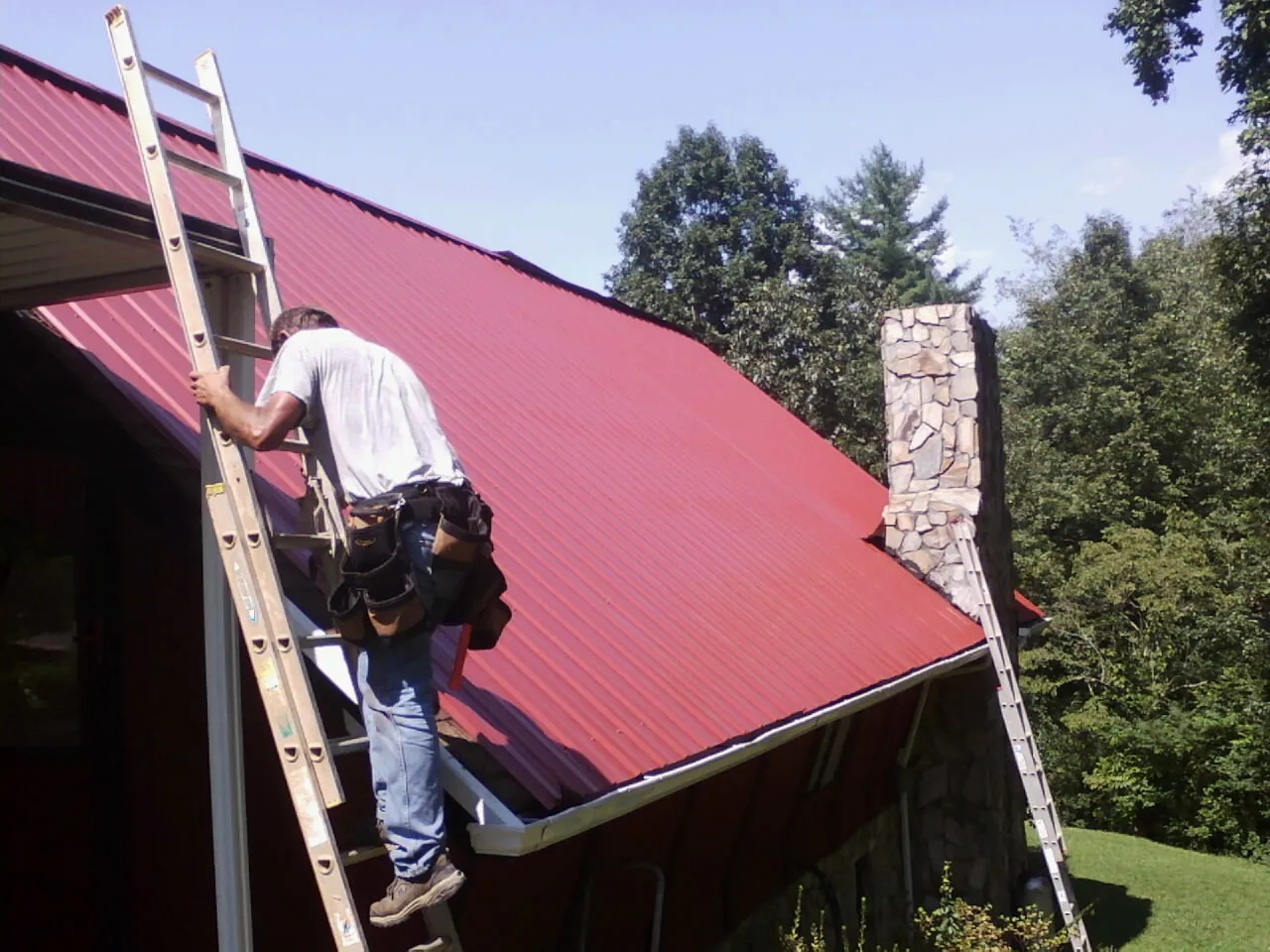 Roofer on a ladder beside a red metal roof and stone chimney — Soffit & Fascia Repair by Roofing Brothers in Port Neches